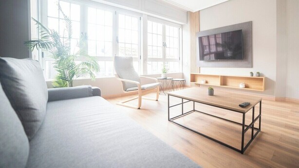 A modern living room with a sofa, a coffee table, a chair, a TV, and a large window with plants.