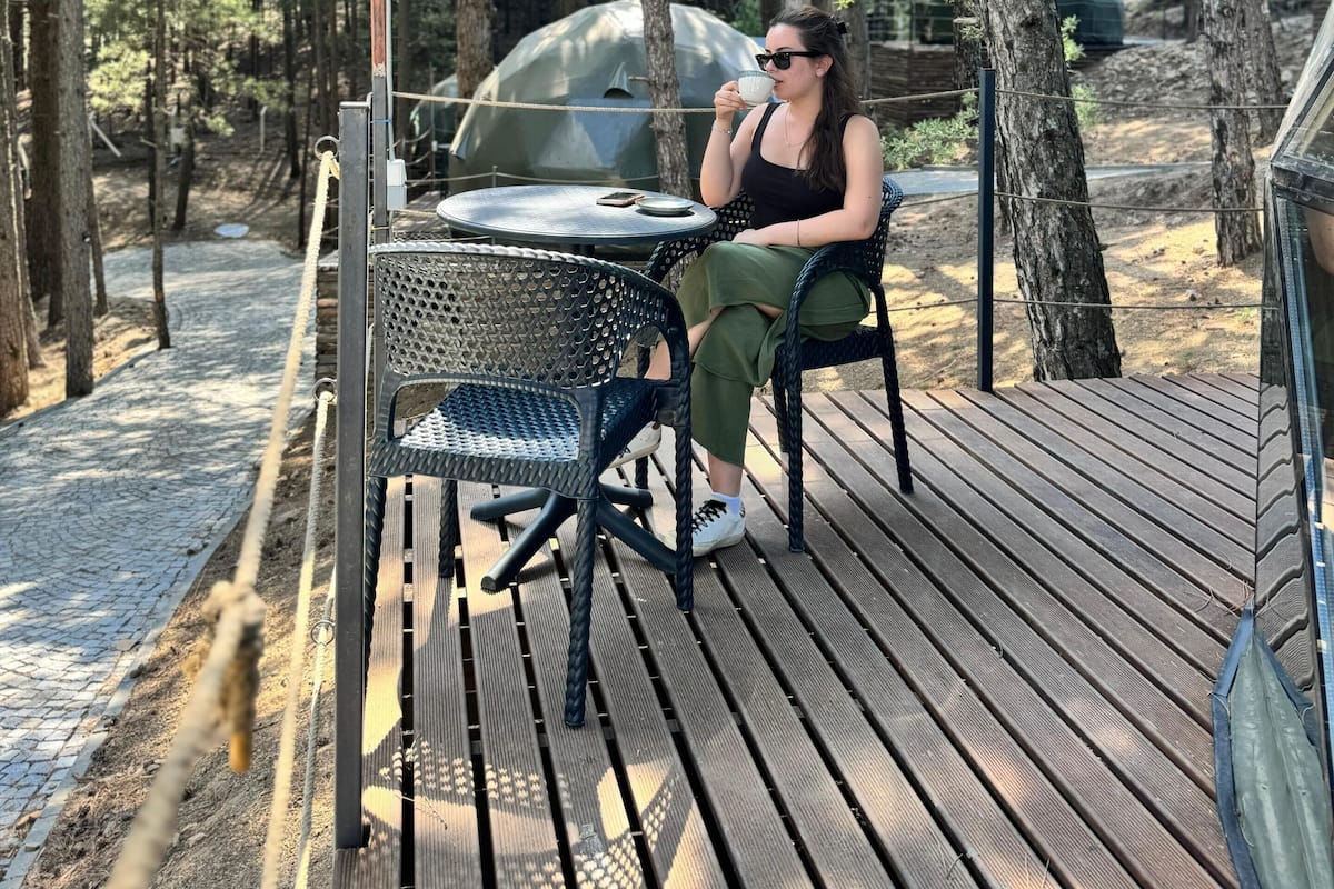 Eine Person sitzt an einem Tisch auf einer Holzdiele in einer bewaldeten Gegend, im Hintergrund ist ein Grill zu sehen.