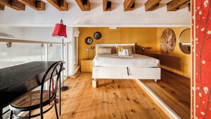 Quarto com teto de madeira, cama com roupa de cama branca, mesa de cabeceira, cadeira e abajur vermelho.