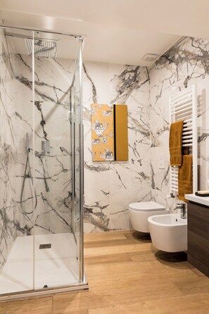 A modern bathroom with a glass shower enclosure, marble walls, a wooden floor, a toilet, a bidet, and a towel rack with towels.