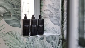 Ikkunan vieressä olevalla hyllyllä on kolme mustaa pulloa, joiden etiketissä lukee: ”Hoitoaine”, ”Suihkusaippua” ja ”Shampoo”. Seinässä on lehtikuviota.