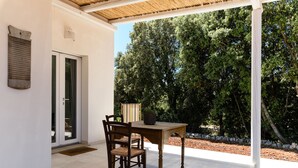 An outdoor patio with a wooden table and chairs, a door with a glass panel, and a view of a wooded area.