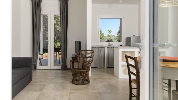 A modern kitchen with a large window, a dining area with a wicker chair, and a sofa in a bright living space.