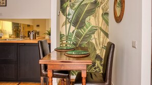 Un comedor con mesa y sillas de madera, una cocina al fondo y un papel tapiz con hojas tropicales.