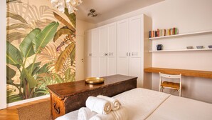 Habitación de hotel con una cama, un aparador de madera, una mesita con un bol, una silla y una estantería con libros y objetos decorativos.