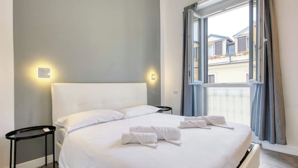 A neatly made bed with white linens and towels, a bedside table, and a window with curtains. - 15min to Duomo, Modern 1 Bedrm Flat A/C, 5G WiFi (Milan)