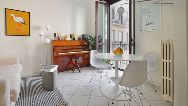 A modern living room with a piano, white chairs, a round table with fruit, and a large window with a balcony view. - Genova25 - One Bedroom Apartment, Sleeps 3 (Milano)