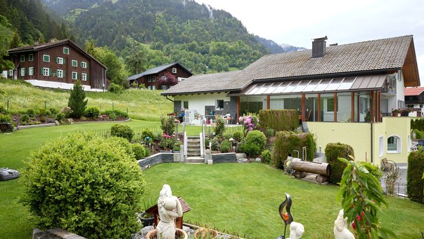 Een huis in de bergen met een verzorgde tuin, een houten schuur en uitzicht op de omliggende bergen.