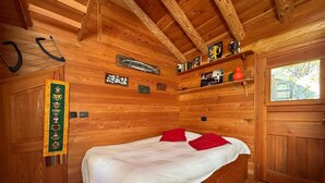 Une chambre dans une cabane en bois, avec un lit, des oreillers rouges, une étagère contenant divers objets et une porte dotée d’une fenêtre.