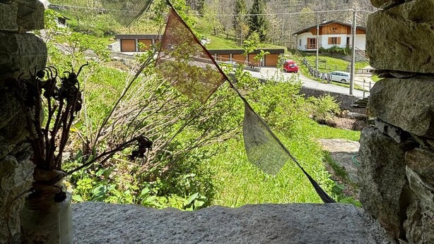 Une fenêtre encadrée de pierres offrant une vue sur un paysage montagneux, une maison en bois et une voiture rouge garée à l’extérieur.