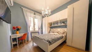 A bedroom with a bed, a desk, and a chair. There is a window with curtains, a chandelier, and a wardrobe. - Chic Modern 2-Bedroom near Metro (Roma)