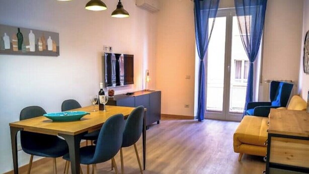 A modern living room with a wooden dining table, blue chairs, a sofa, and a TV. - Chic Modern 2-Bedroom near Metro (Roma)