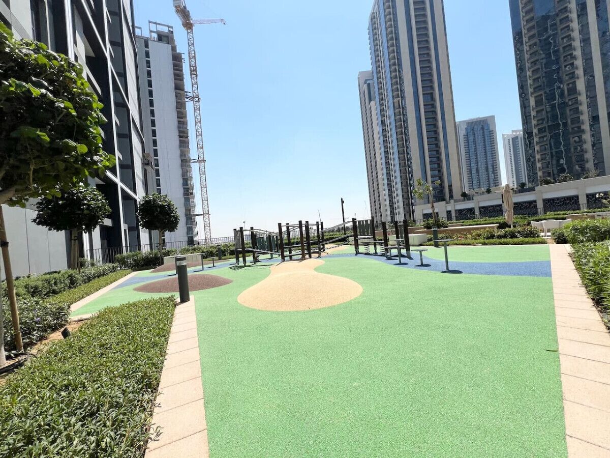 Un parque infantil con césped verde y un arenero, rodeado de edificios altos y una grúa de construcción.
