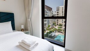A hotel room with a large window, a neatly made bed, a bedside lamp, and a view of a swimming pool and buildings.