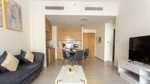 Una sala de estar moderna con un sofá, una mesa de centro y un televisor. Se observa una cocina al fondo, con armarios de madera, y un comedor con sillas.