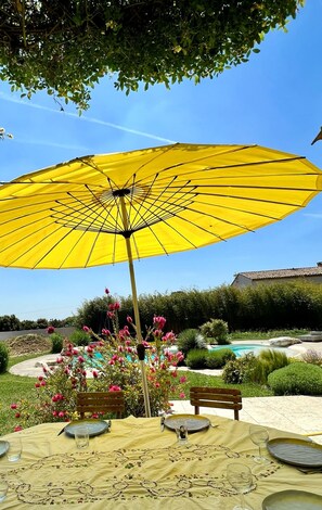 Un espace repas agrémenté d’un parasol jaune, d’une piscine et d’un jardin où fleurissent des plantes aux couleurs roses.