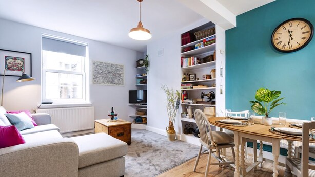 Una sala de estar acogedora con un sofá, una mesa de comedor para dos personas, una estantería y un reloj de pared grande.