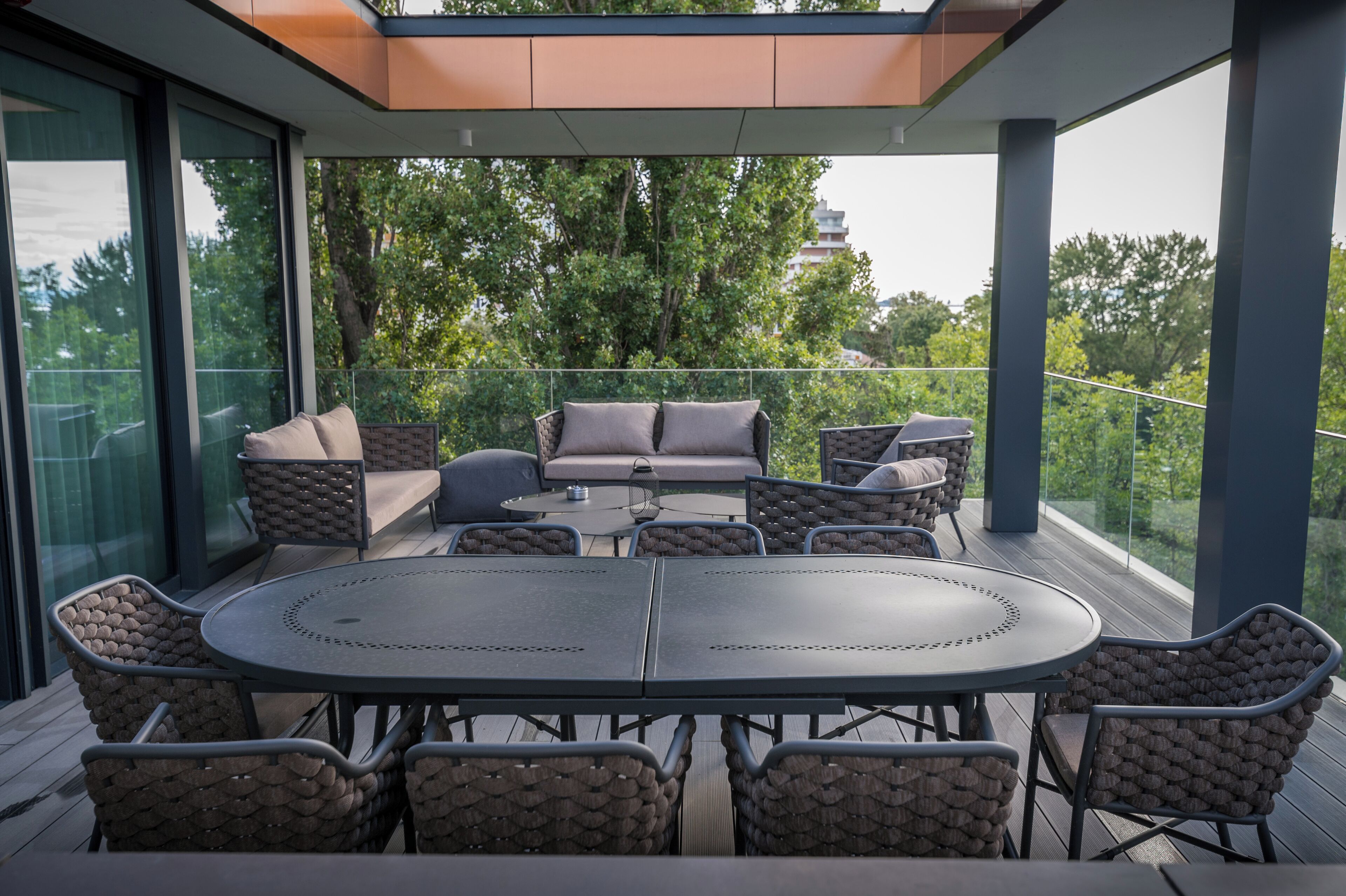 A modern outdoor patio with a round table and wicker chairs, surrounded by glass walls and a view of trees and a distant building.