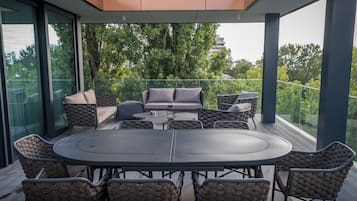 A modern outdoor patio with a round table and wicker chairs, surrounded by glass walls and a view of trees and a distant building.