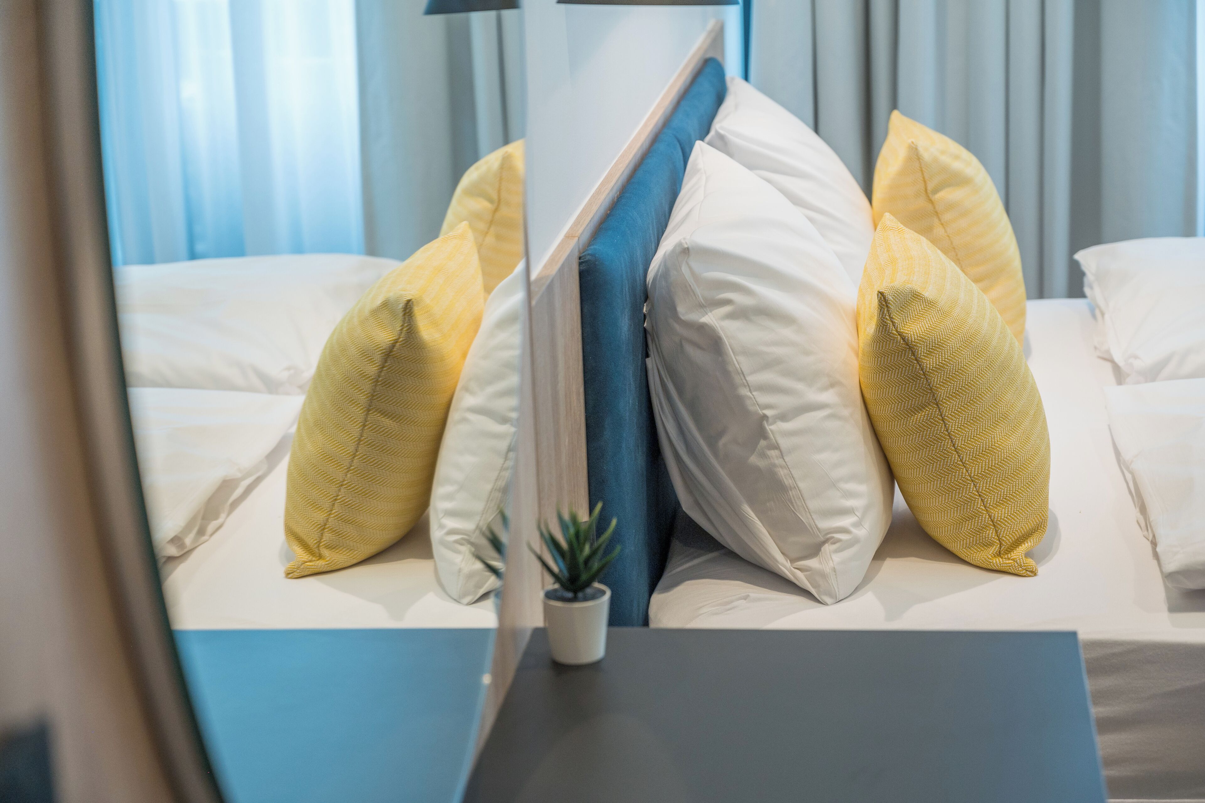 Een keurig opgemaakt bed met witte en gele kussens, een kleine potplant en een blauw hoofdeinde.
