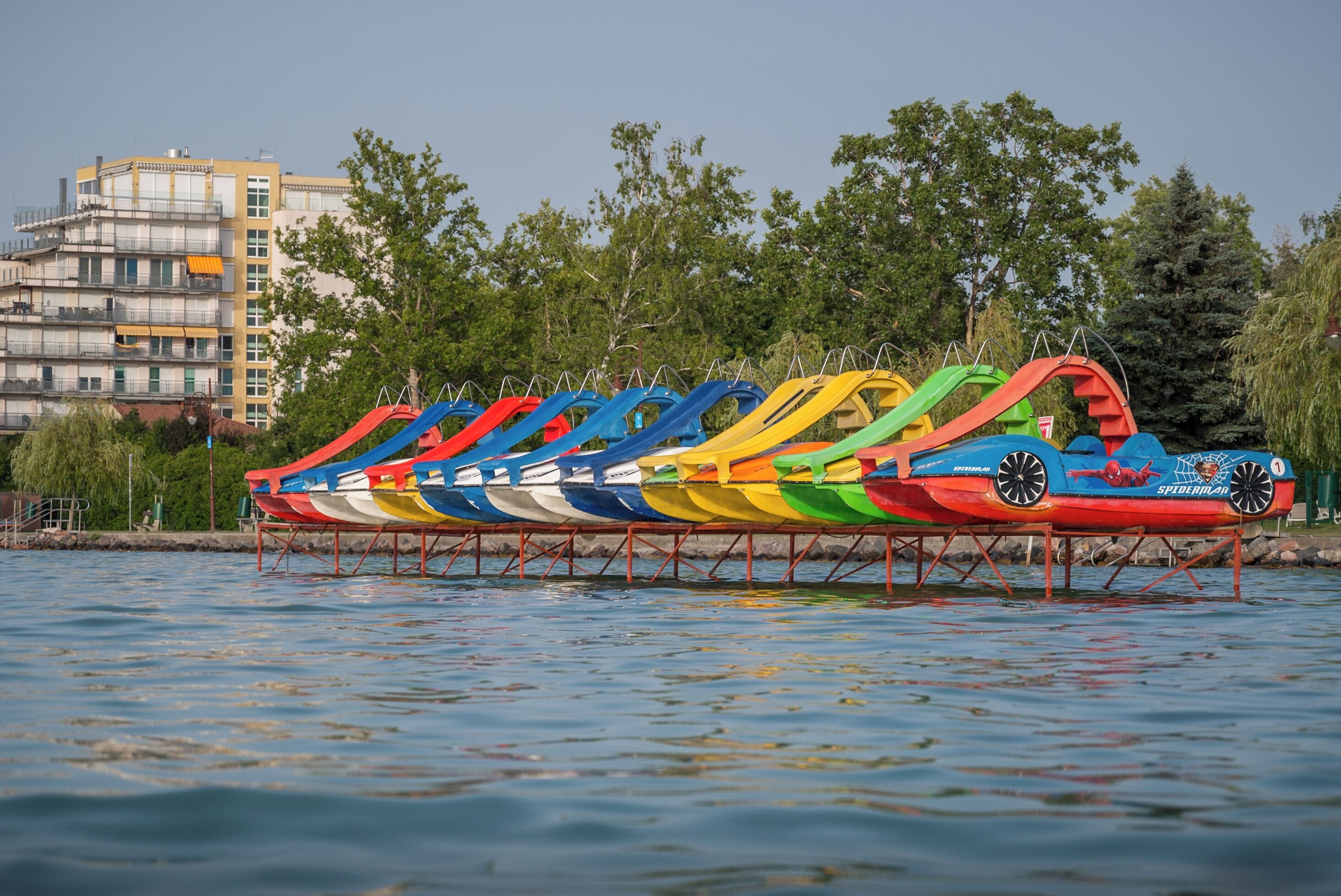 A row of colorful inflatable boats on a body of water with a building and trees in the background.