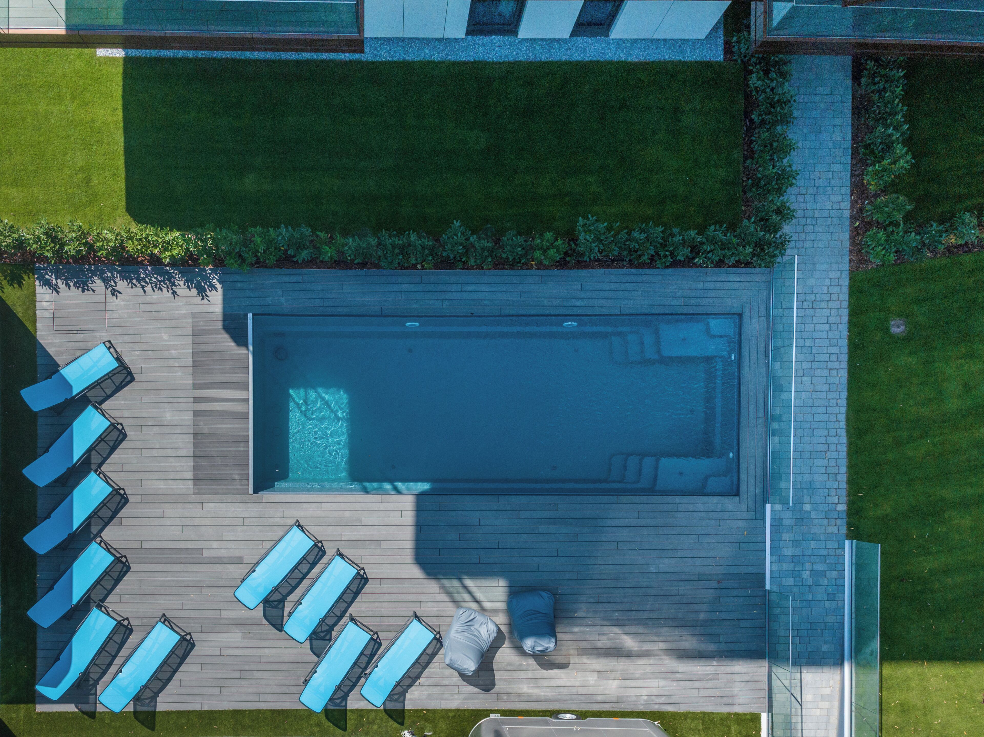Vue aérienne d’une piscine rectangulaire dotée d’une terrasse en bois, entourée de chaises longues et d’un petit espace salon.
