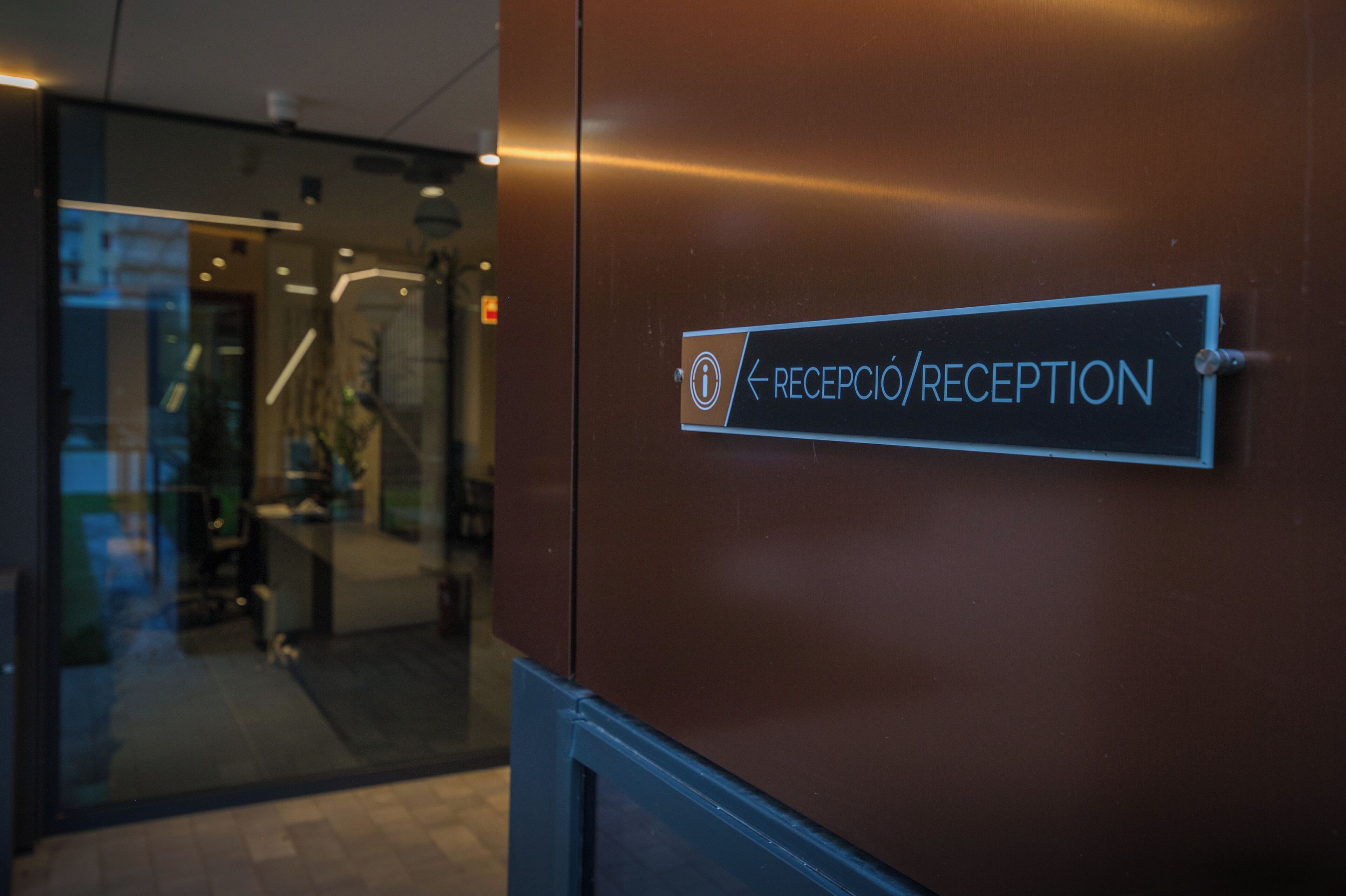 En skylt med text för receptionen, placerad på en brun vägg, med en glasdörr synlig i bakgrunden.