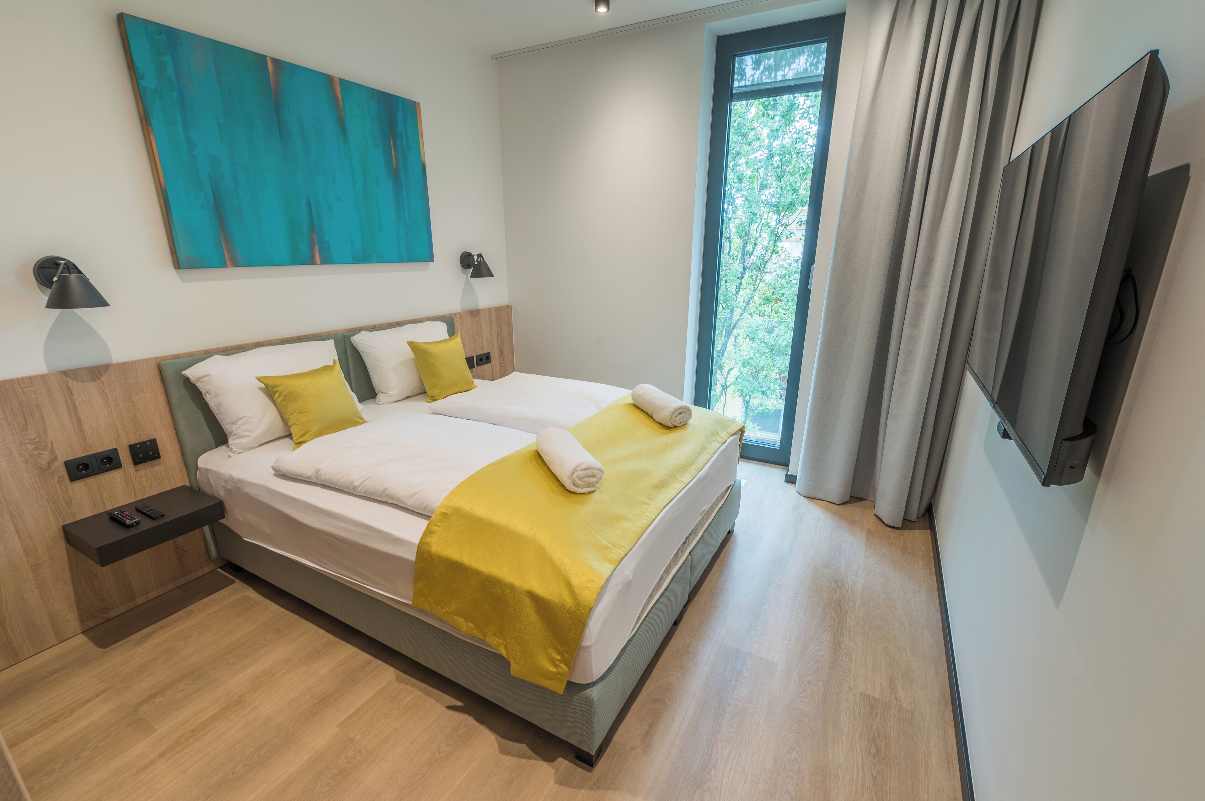 A modern hotel room with a large bed, bedside tables, a TV, and a window with curtains.