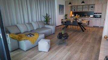 Un salon moderne avec un canapé gris, un petit pouf blanc et une table basse noire. On y trouve également un coin repas avec une table et des chaises en bois, ainsi qu’une cuisine dotée de meubles en bois et d’une suspension noire.