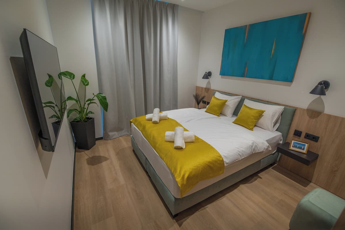 A modern hotel room with a bed, a flat-screen TV, a potted plant, and a painting on the wall.