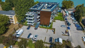A modern multi-story hotel with a rooftop terrace, parking area, and a view of the lake. - BJ81 Suite Hotel (Siófok)
