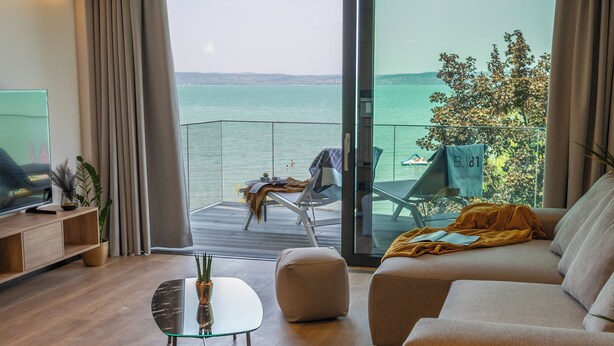 A modern living room with a sofa, ottoman, and glass coffee table. A balcony with a view of the sea and a flag indicating a hotel suite is visible. - BJ81 Suite Hotel (Siófok)