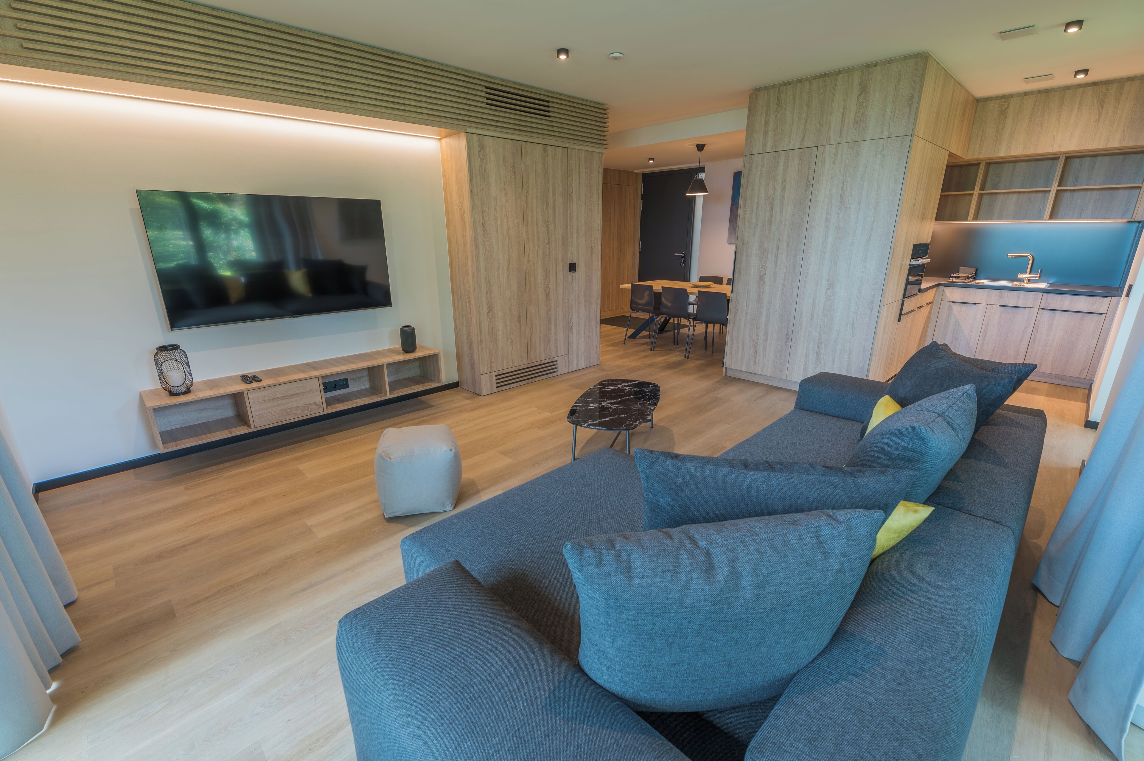 A modern living room with a large flat-screen TV, grey sofas, a coffee table, and a kitchen area in the background.