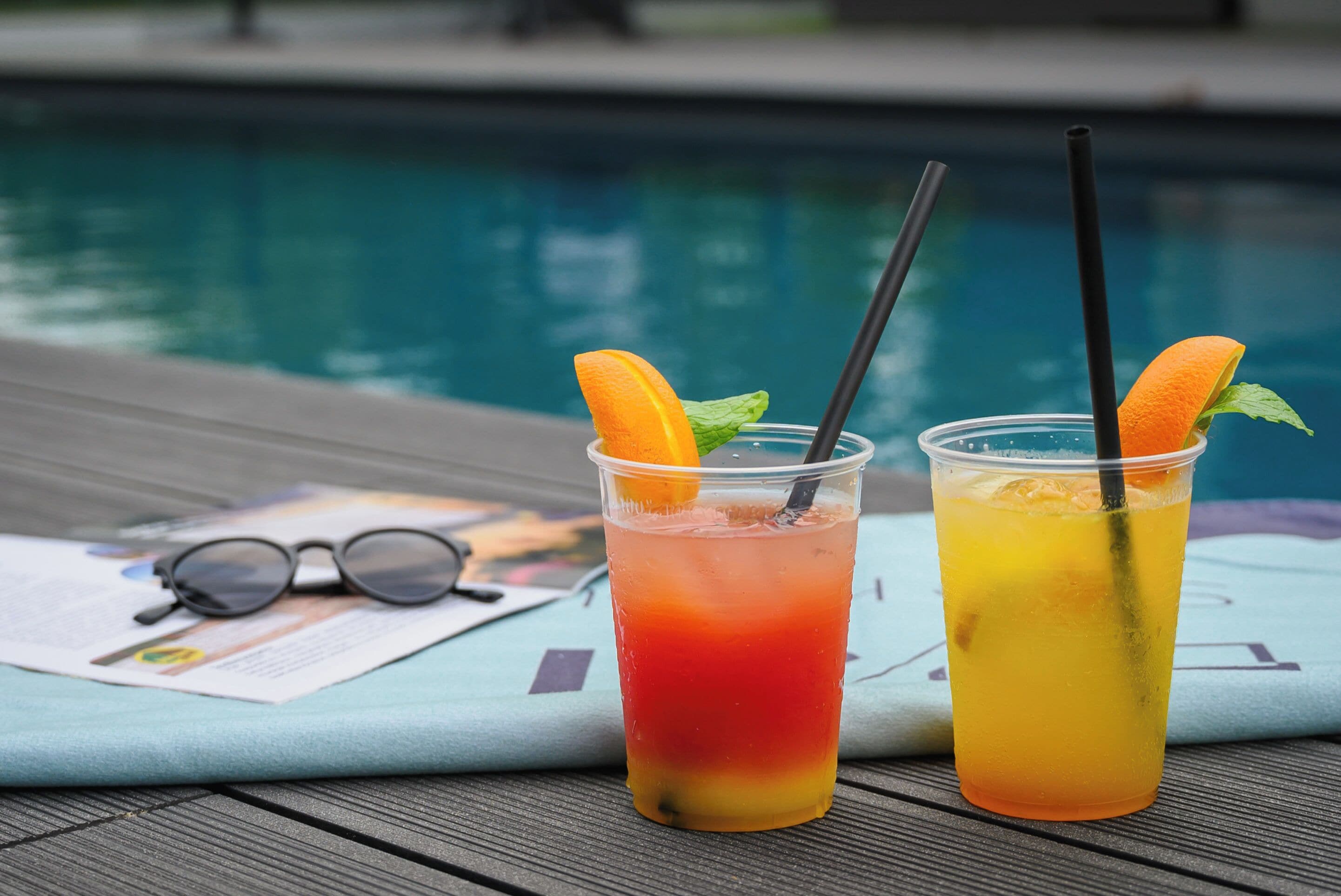 Two drinks with straws and orange slices on a table near a pool.