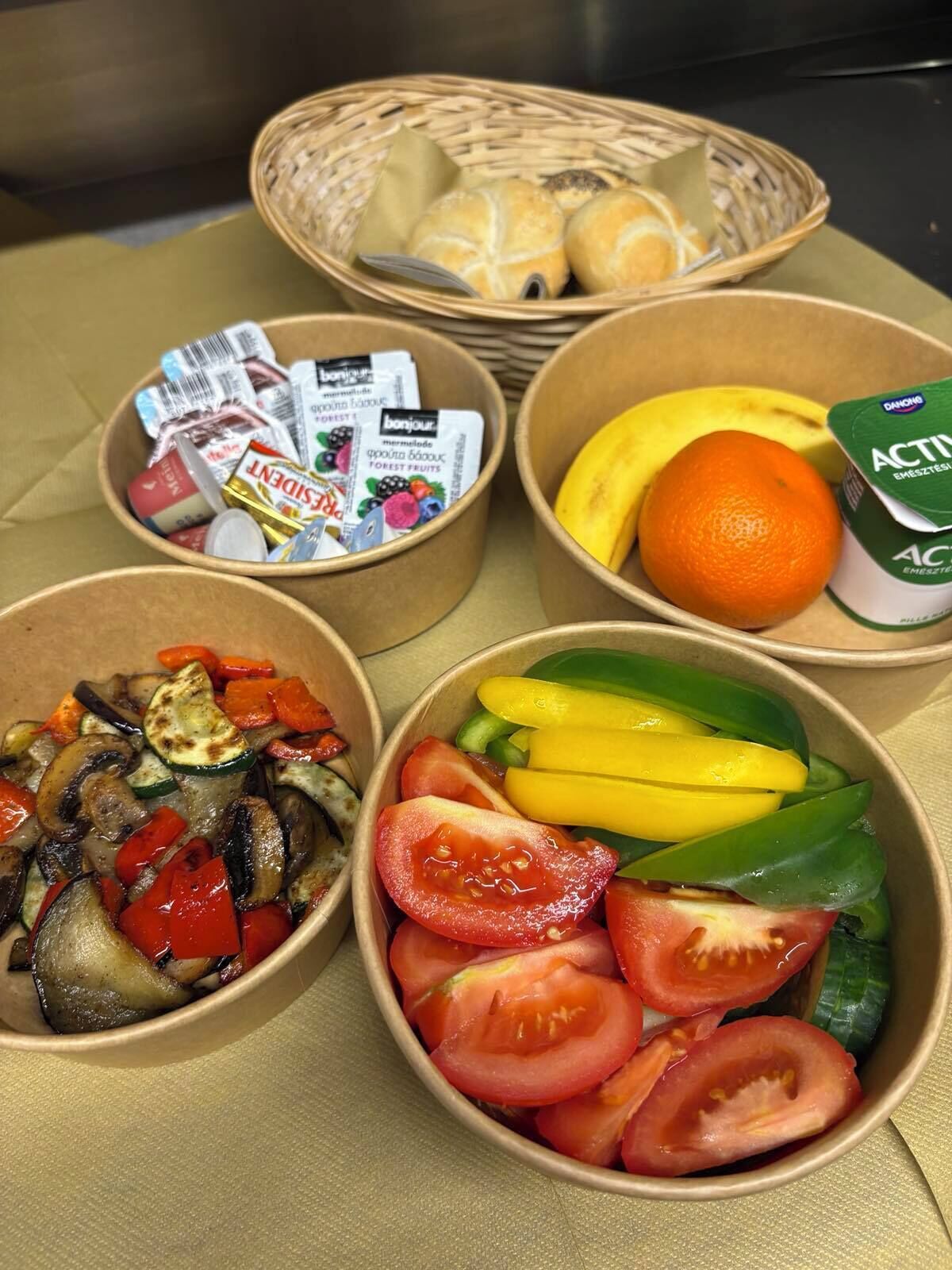 Una comida dispuesta con platos de verduras, una cesta de pan y un cartón de jugo de naranja.