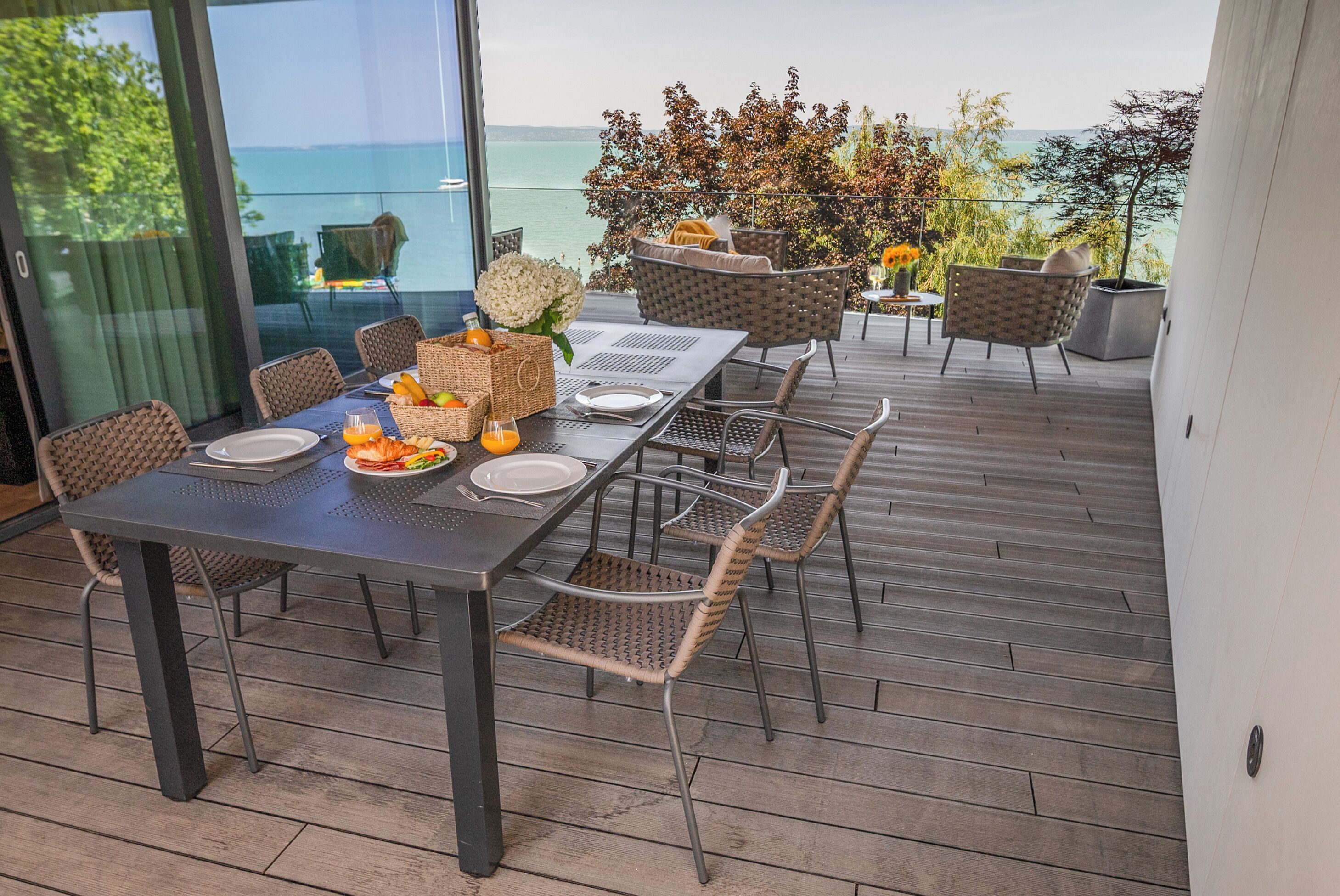 A dining area with a table set for six, surrounded by wicker chairs, overlooking a scenic view of the sea.