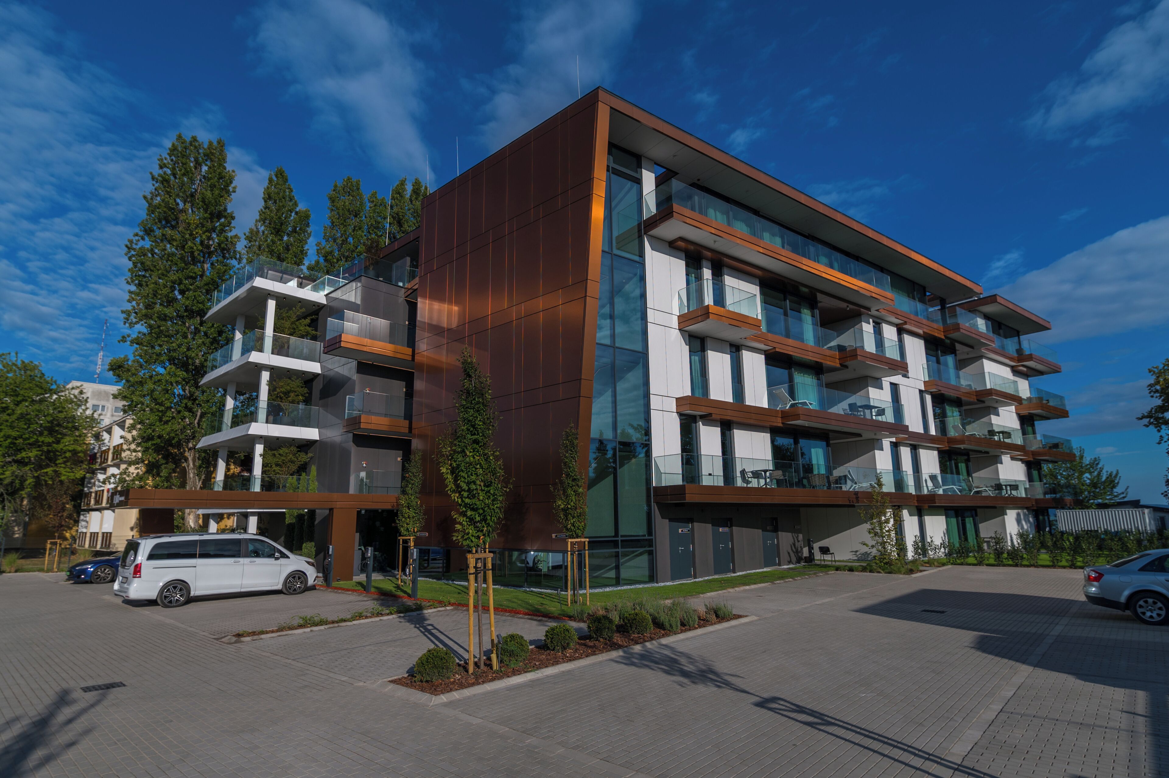 A modern multi-story building with balconies, a parking area, and surrounding greenery.
