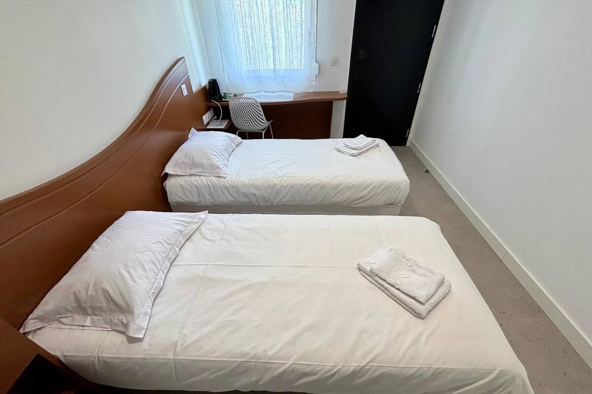 Ein Hotelzimmer mit zwei Einzelbetten, einem Schreibtisch mit Stuhl und einem Fenster mit leichtem Vorhang.