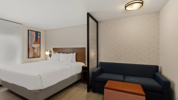 A hotel room with a bed, a sofa, a small table, a nightstand, a wall-mounted lamp, and a ceiling light.