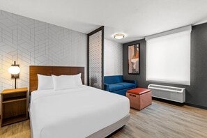 A hotel room with a large bed, a nightstand, a blue chair, a red ottoman, a wall-mounted heater, and a geometric-patterned wall.