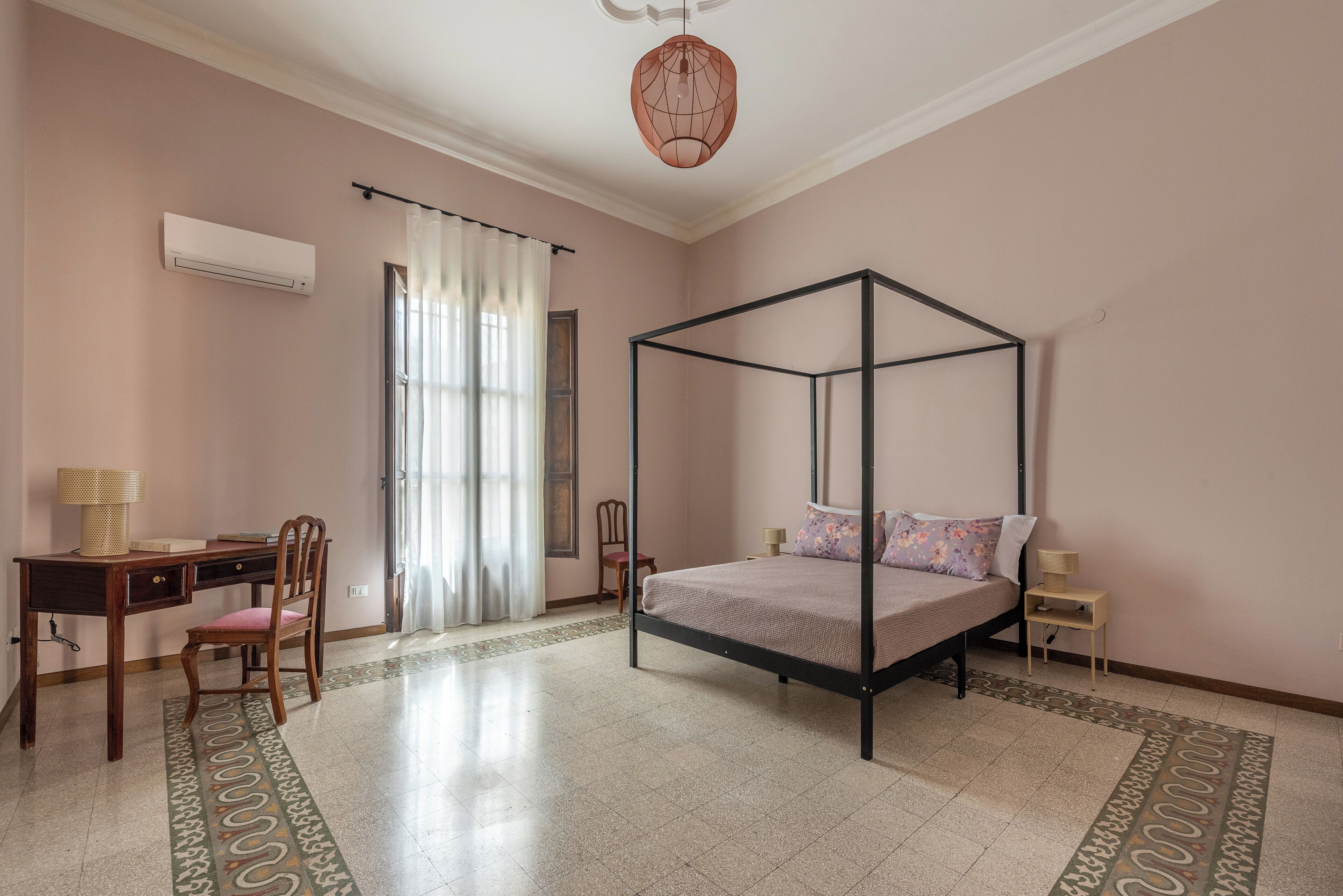 A bedroom with a four-poster bed, a desk with a chair, a small bedside table, and a window with curtains.