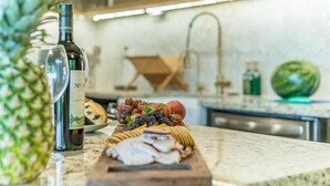 A kitchen counter with a bottle of wine, a wooden board with various foods, a pineapple, and a watermelon.