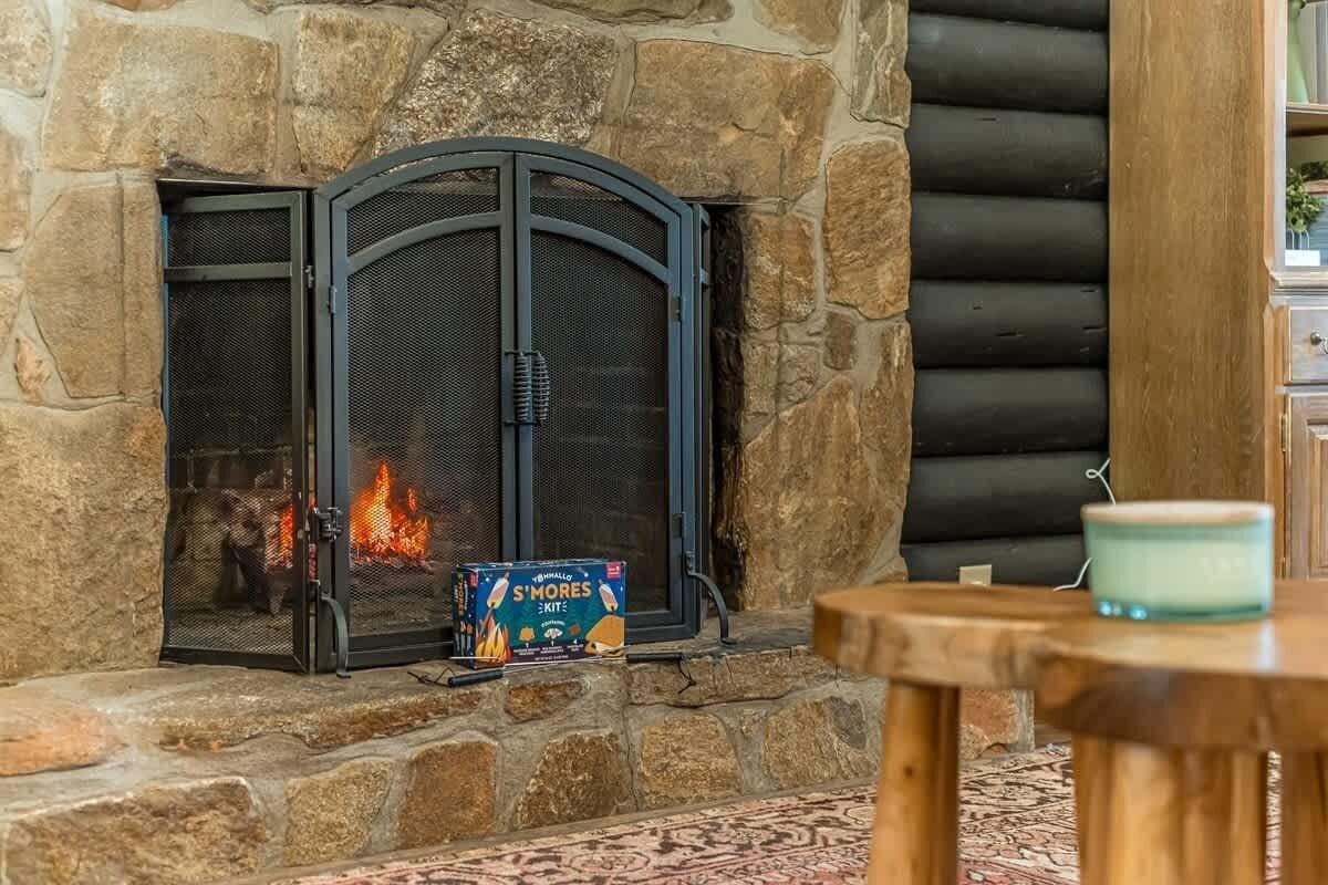 A stone fireplace with a metal screen door, a box of S'mores kits, and a teal candle on a wooden table.