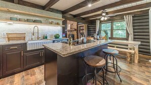 A log cabin interior with a kitchen island, bar stools, wooden beams, and a window with curtains.