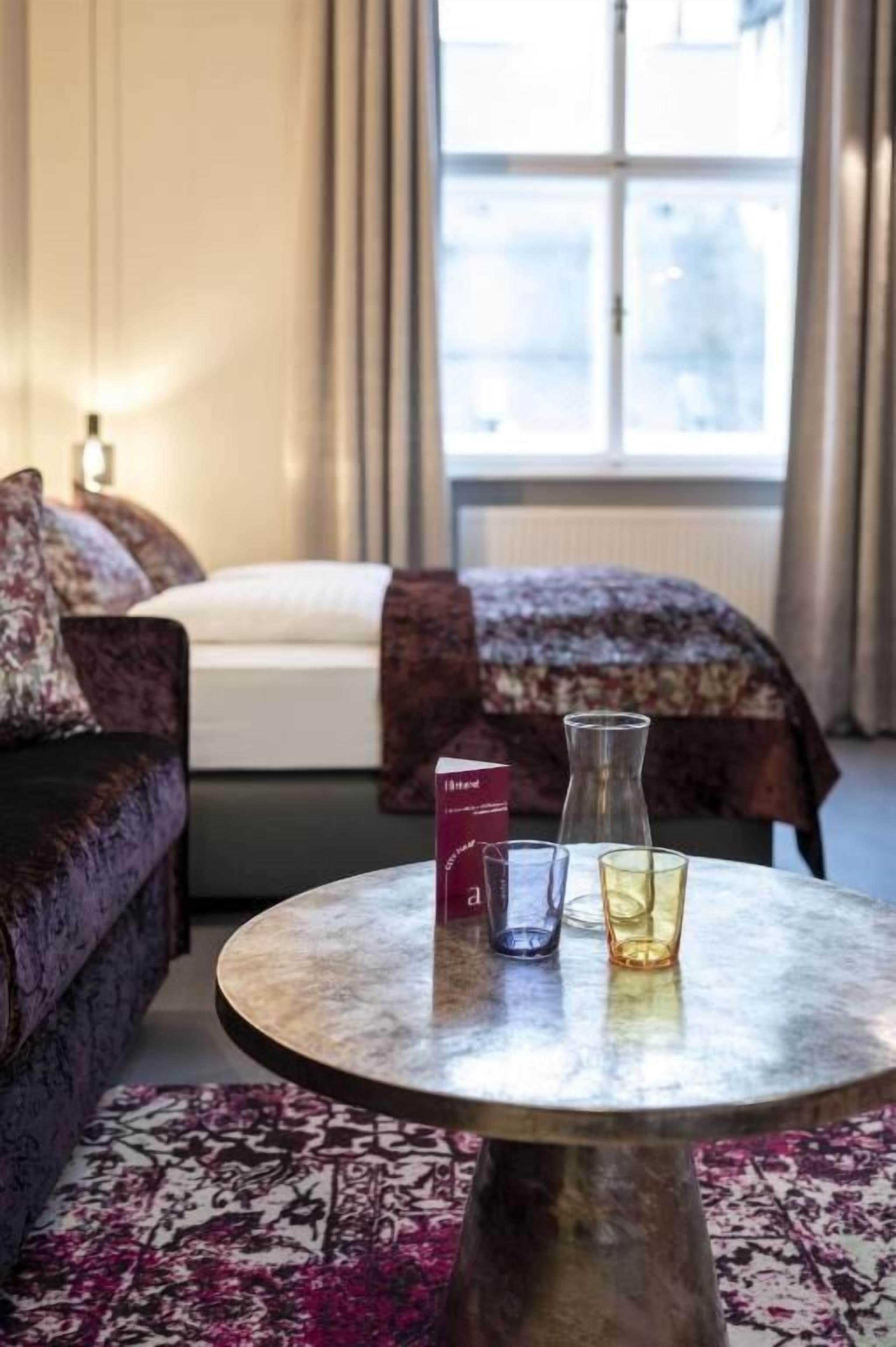 Ein Hotelzimmer mit Bett, einer Couch, einem runden Tisch mit Buch und Gläsern sowie einem Fenster mit Vorhängen.