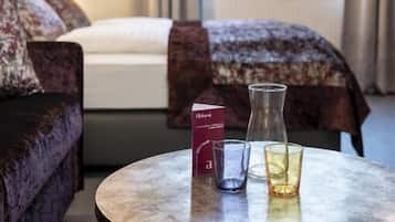 Ein Hotelzimmer mit Bett, einer Couch, einem runden Tisch mit Buch und Gläsern sowie einem Fenster mit Vorhängen.