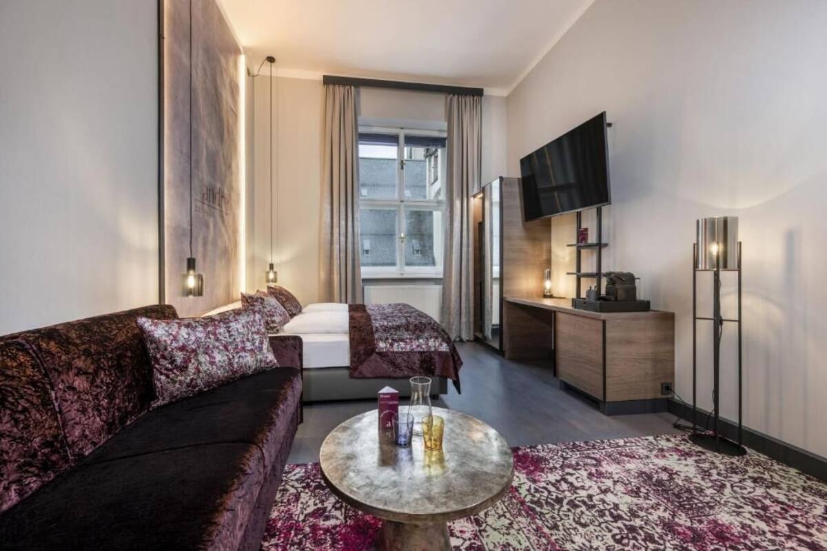 A modern hotel room with a sofa, a round coffee table, a bed, a TV, and a desk.