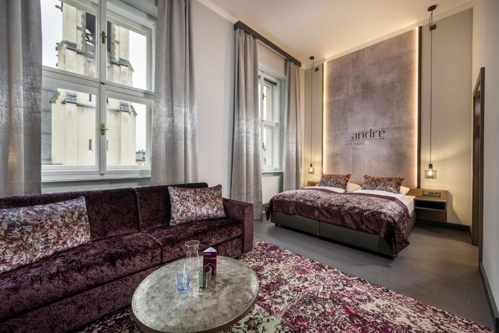A modern hotel room with a sofa, a round coffee table, a bed with a headboard, and a large window with curtains.