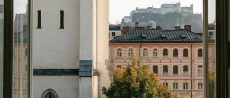 A window with a view of a building with a clock tower and another building with a red facade.