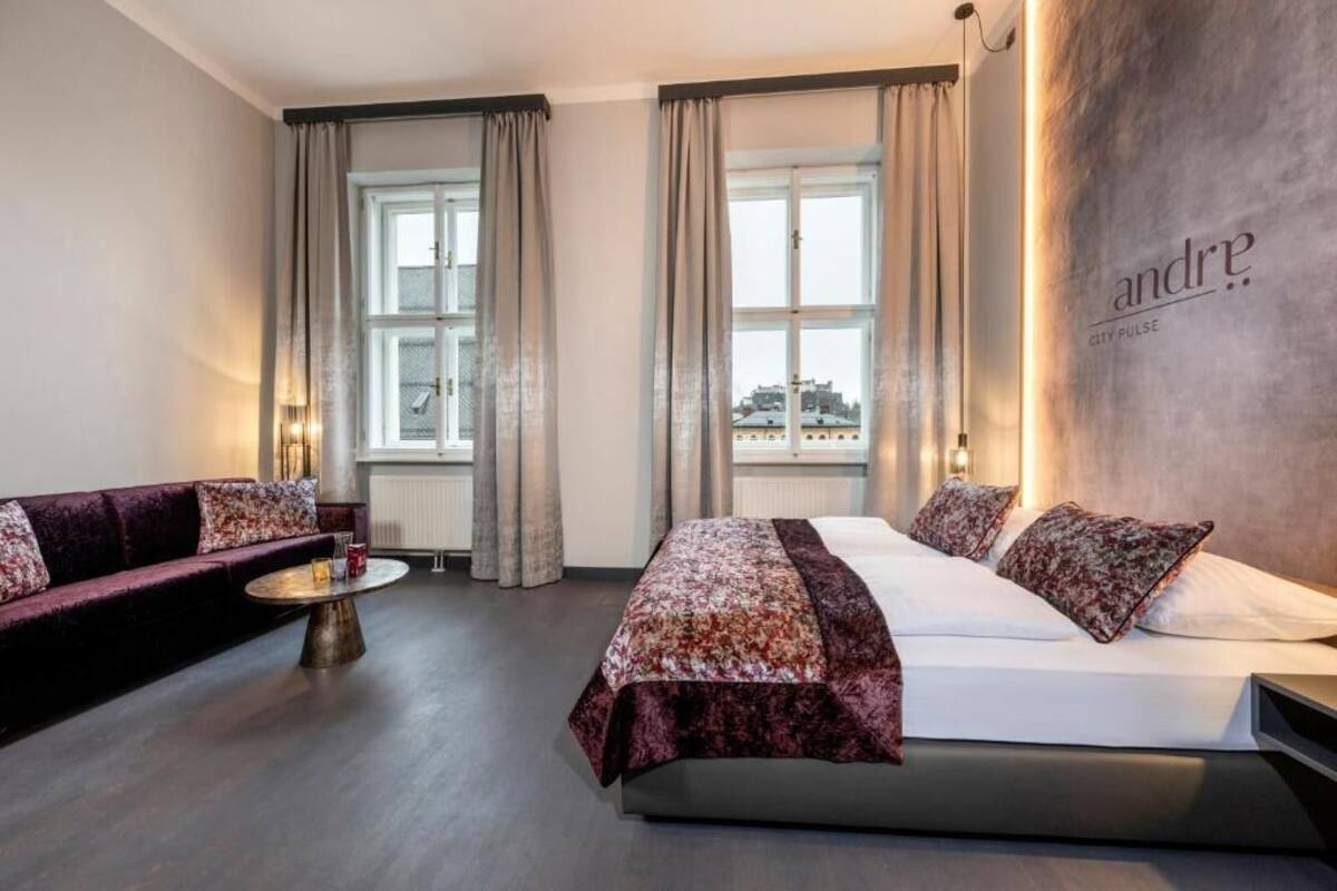A hotel room with a bed, a sofa, a round table, and large windows with curtains.